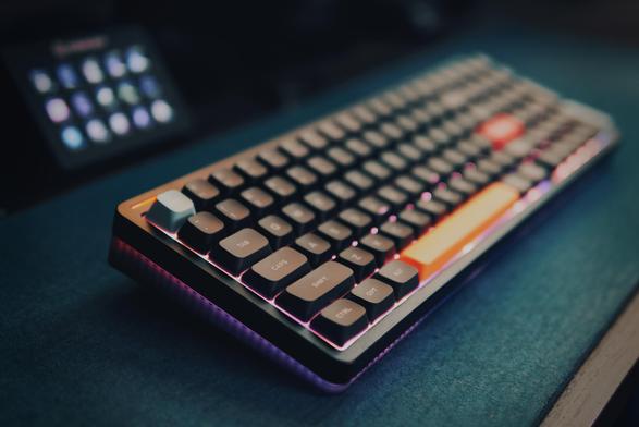 A picture of my desk. A black keyboard with RGB lighting is resting on a teal felt mat. in the background stream deck is visible.