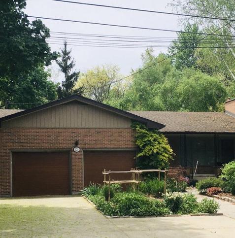 A single-story brick house with a brown roof and two garage doors. One half of the driveway has been dug up to create a front garden that features a variety of plants and flowers, along with a wooden structure. Surrounding the house are trees and greenery, and overhead power lines are visible.