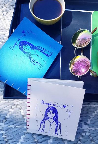 Sur un plateau noir, un carnet bleu, des lunettes de soleil rondes dans les verres desquelles se reflète un arbre en fleur et le soleil. Un autre carnet, blanc, est posé contre le plateau. Carnets à reliure copte et linogravure montrant une femme fermant les yeux. Au dessus d'elle l'inscription "Respire... ♡"