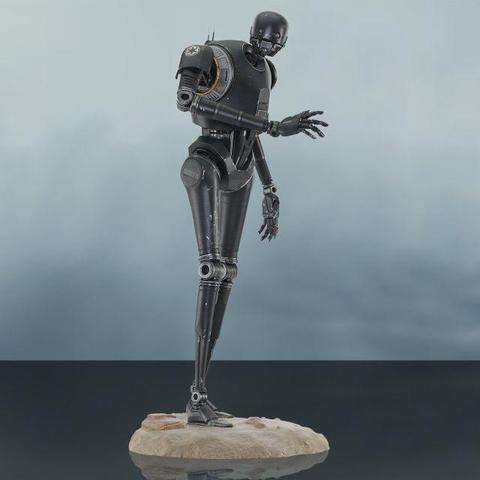 Statue de K-2SO sur un socle ressemblant à du sable. Son bras droit est plié vers le haut, alors que le gauche pend. Le droïde penche légèrement vers l'avant. Le fond de la photo est dans des teintes de gris.