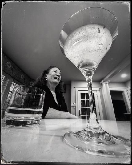 Low angle black and white photo looking up at a martini glass with a smiling woman in the background.