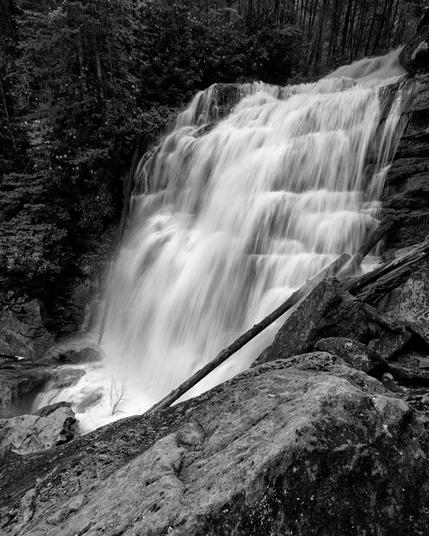 A black and white image of a waterfall centered in the frame with a large boulder in the foreground and trees framing the top and left of the frame.