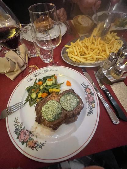 A plate of steak topped with green herb butter is served alongside mixed sautéed vegetables and a portion of French fries. Several glasses of red wine and water are on the table, which is adorned with a floral-patterned. 