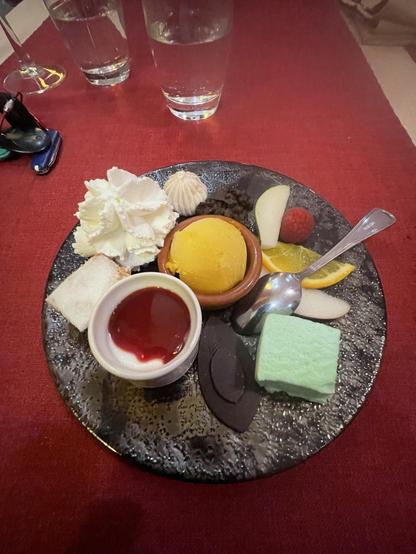 A dessert plate featuring a scoop of orange sorbet in a ceramic dish, accompanied by cream, assorted fruits (including apple, raspberry, and orange), a small cup of red sauce, chocolate pieces, and a light green sweet.