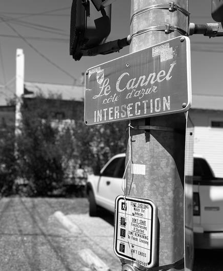 Light post, up close, in black and white, with a sign that explains the different walk signs. Above it is a sign that says “Le Cannet, Côte d'Azur intersection.” A house and a truck are in the background. 
