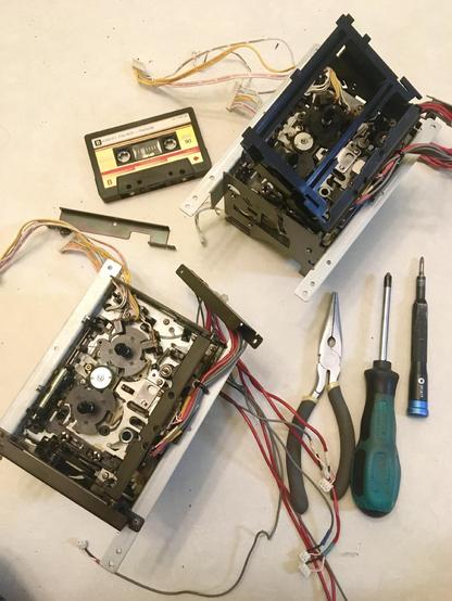 Two tape mechanisms from Akai MG614 multitrack cassette recorders have been removed from their respective machines and rest on the floor alongside a cassette tape, some pliers and two screwdrivers. One of the mechanisms is missing its door.