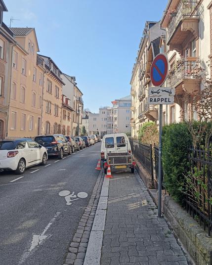 Une camionnette type Citroen C15 avec une remorque, stationnée sur le trottoir et bloquant intégralement le passage des piétons. Les deux roues gauche de la camionnette sont dans le caniveau sur la chaussée. Le conducteur a disposé deux cônes orange et blanc derrière la remorque et sur la chaussée au coin arrière droit de sa voiture. La rue est déserte.