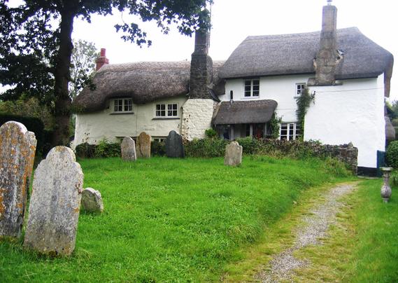 In the foreground is a churchyard with several headstones, lots of green grass and a tree. On the edge, in the background, are two cottages painted white with thatched roofs and very tall chimneys.