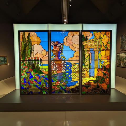 Hyper saturated and colorful stained glass artwork in 3 panels showing an outdoor scene with trees and roman columns. 
