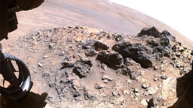 An image captured by the left front hazard avoidance camera of the Mars rover Perseverance, featuring a rocky mound and a wheel of the rover, with a Martian landscape in the background. 