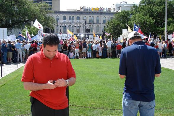 Photograph of two young men, one with a red shirt facing us, the other with a blue shirt with his back to us. Each are working on their cell phones. In the distance is a crowd of taxi protesters at the first "Tea Party" rally.
