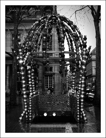 The image captures a striking installation made up of shiny metallic elements, resembling a whimsical structure that draws the eye with its intricate design. It features a dome-like shape comprised of numerous reflective components, creating a sense of movement as they arch over a central area. The ground below is wet, likely from recent rain, which enhances the reflections of the metallic pieces, adding a layer of depth and texture to the scene.
In the backdrop, there are buildings that suggest an urban environment, their architecture adding context to the installation. The interplay of light and shadow in black and white emphasizes the contours and details of the metallic structure, evoking a sense of both modernity and artistic expression.
This piece could be interpreted as a commentary on urban life, blending nature with man-made materials, or it might represent a sanctuary amidst the bustling city. The composition invites viewers to pause and reflect, encouraging a moment of contemplation within the busy atmosphere surrounding it. Overall, it embodies a blend of creativity, design, and the interplay between the natural and the artificial.