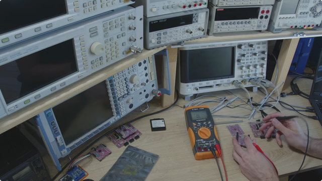 Screen capture: person probing a small board on a desk. On the stands in the back, multiple layers of measurement devices.