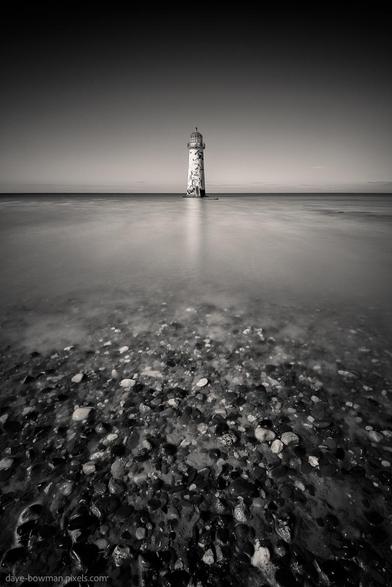 A monochrome photograph of Talacre Lighthouse, also known as Point of Ayr Lighthouse, centred on the horizon. In the foreground, the receding tide reveals a pebbled beach and smooth sand in the shallow water. Above, the clear, cloudless sky emphasises the lighthouse’s solitary presence on the North Wales coast, creating a simple yet striking composition.