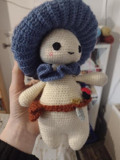 A doll sized, crocheted mushroom person, with a pale cream body, blue neck frill and cap gill. They have a little brown tool belt with a bag, spade, and knife attached.