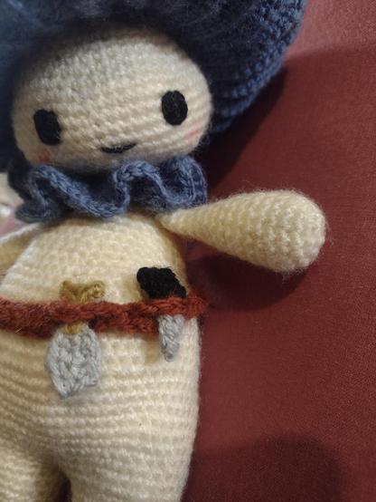 A doll sized, crocheted mushroom person, with a pale cream body, blue neck frill and cap gill. They have a little brown tool belt with a bag, spade, and knife attached. The view is a close-up of the belt, with the mushroom person looking down at it.