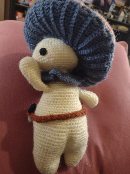 A doll sized, crocheted mushroom person, with a pale cream body, blue neck frill and cap gill. They have a little brown tool belt with a bag, spade, and knife attached. The view is from the back, showing a distinct little bottom. The mushroom person has their hand up to their mouth in a cheeky pose.