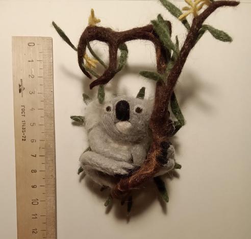 On a white background lies a craft in the form of a koala. To the left of it lies a ruler, by which you can see the size of the craft. About 12 centimeters.
Brooch made of nettle fibres by dry felting, in the form of a koala. The koala sits on a branch of a blossoming eucalyptus and holds onto it with one front and one back paw. It is resting.
Thank you for your attention!