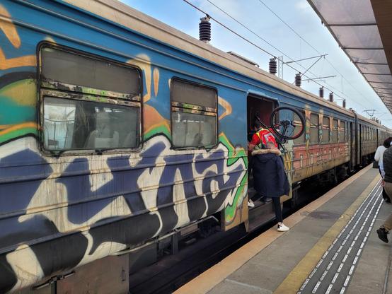 Old train covered in graffiti (windows included) with a mountain biker bording with his bike on the shoulder.
