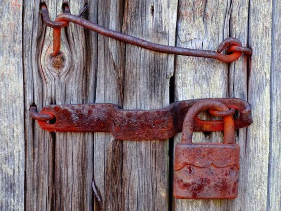 Detailaufnahme einer alten Scheunentür mit rostigen Riegel und Vorhängeschloss sowie einfachem Hakenriegel. Farben: altes verwittertes Holz und rotbrauner Rost.
