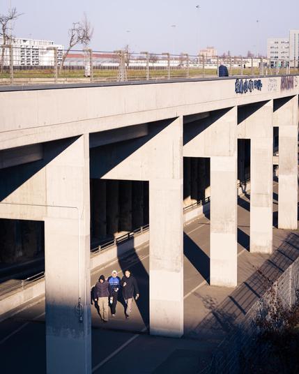 A group of three people walks under a large concrete overpass, bathed in strong sunlight that creates dramatic shadows. The massive pillars of the structure form a repetitive pattern, contrasting with the open sky and modern buildings in the background. A lone pedestrian walks along the top of the overpass, adding a sense of scale and depth to the urban scene. The atmosphere is quiet and contemplative, with the interplay of light and shadow emphasizing the geometric forms of the architecture.