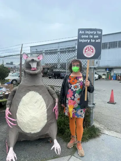Urban Ore worker stands outside gate entrance to car parks. They are standing next to “Scabby the Rat”  puppet.
A sign says “An injury to one is an injury to all” - IWW Industrial Workers of the World 
