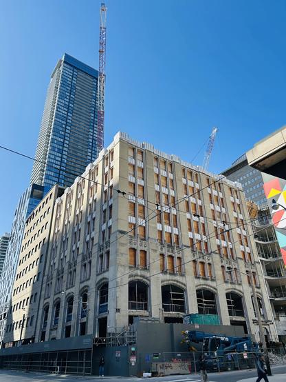 An image of a construction site featuring a partially renovated building with boarded windows and a crane above. A modern high-rise towers in the background under a clear blue sky. The foreground shows construction equipment and workers, alongside a construction barrier.