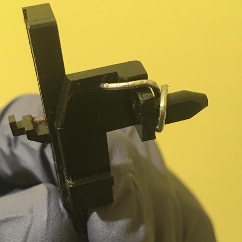 A paperclip is bent through and around a Slide Eject black plastic part which has been removed from the tape mechanism of an Akai MG614 multitrack cassette recorder. A gloved hand is holding the part in front of a yellow wall.