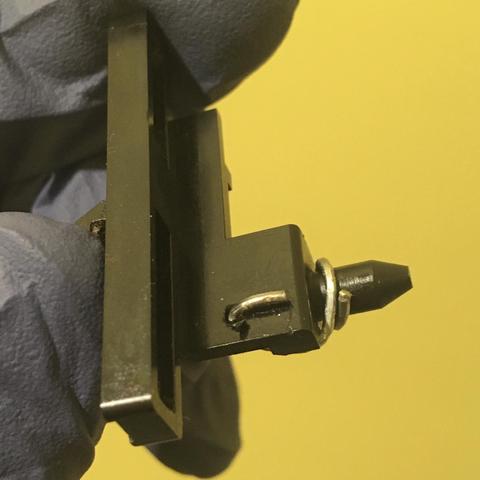 A paperclip is bent through and around a Slide Eject black plastic part which has been removed from the tape mechanism of an Akai MG614 multitrack cassette recorder. A gloved hand is holding the part in front of a yellow wall.