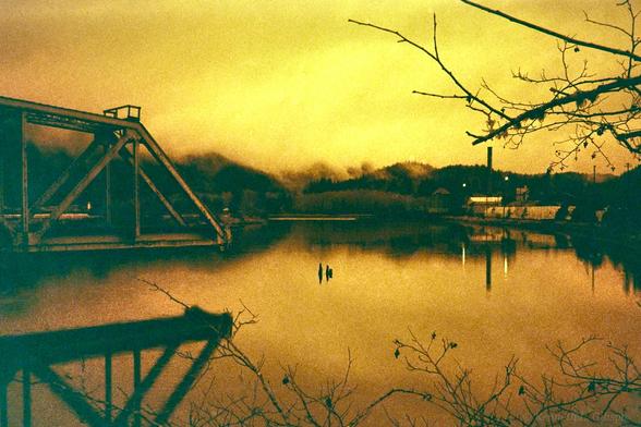 Redscale image of trestle over water with lumber mill in background.