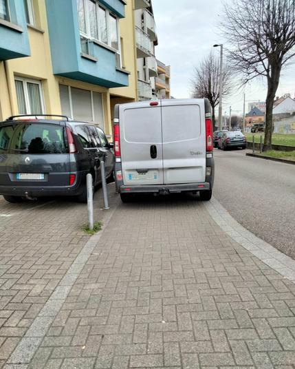 Une camionnette grise stationnée sur le trottoir, à côté d’un Renault Espace lui bien garé dans un parking privé. Les deux formes une sorte de mur rendant la vue impossible pour les piétons. De l’autre côté de la rue, 5 mètres devant, une place de stationnement libre et gratuite.