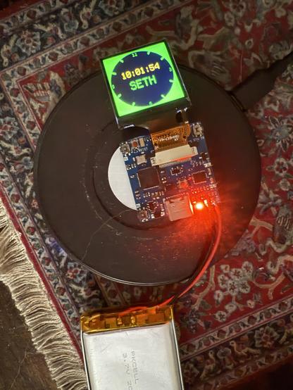 Pictured is the RP Watchy, an RP2040 powered watch project with an RTC from Abracon, an IMU from STM, a 240x240 display, and Qi wireless charger from Texas Instruments. The RP Watchy has an amber LED illuminated to show that it is actively charging via the Qi charger it is sitting on.
