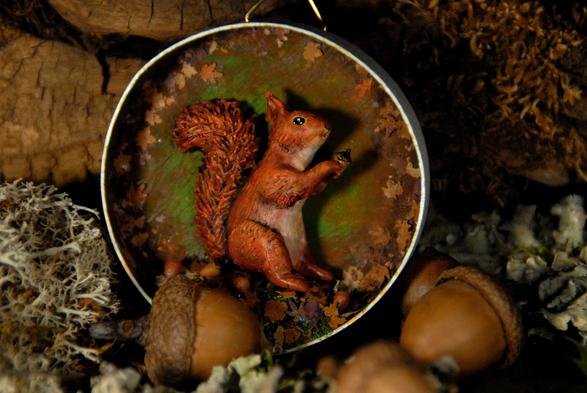 Diorama dans un boite ronde en bois contenant un petit écureuil qui tient un gland dans ses pattes avant. La boite est entourée d'éléments forestiers : lichen, feuilles mortes, glands