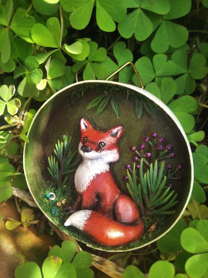 Diorama dans un boite ronde en bois contenant un petit renard assis au milieu de la végétation. Des petits trésors sont visibles à ses pieds : pièces, anneau, graine. La boite est entourée de feuilles d'oxalis