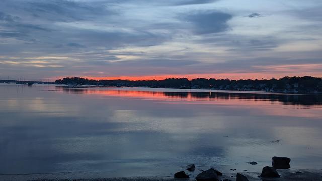 Historical Documentation in the form of a photograph of a rising sun over the ocean bay at the Historic Pioneer Village in Forest River Park, Salem, MA. The sky is filled with vibrant colors of orange, pink, and purple, with the sun rising in the distant background. The water is calm and reflects the colors of the sky, creating a mirror-like effect. On the right side of the image, there is a rocky shoreline with a few rocks scattered across it. In the distance, there are trees and buildings silhouetted against the sky. The overall mood of the photograph is peaceful and serene.