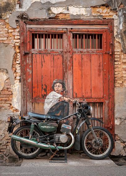 A photograph of a piece of street art in George Town, Penang, Malaysia, where a painted young boy on a rustic red wooden door appears to sit on a real motorbike parked against the wall. The artwork seamlessly blends with the physical elements, making it look as though the boy is ready to ride. The weathered textures of the door and bike add character to the scene, creating a playful yet nostalgic effect.
