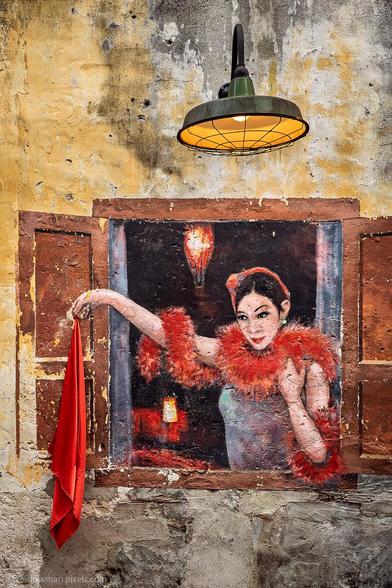 A photograph of a vibrant mural in Kuala Lumpur’s Kwai Chai Hong, a historic corner of Chinatown near Petaling Street. The artwork depicts a lady waving a red scarf, her pose adding a sense of movement to the scene. Above the mural, a vintage lamp casts a warm glow, highlighting the rich colours and intricate details of the painting against the textured wall.