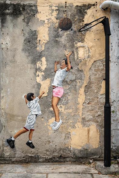 A photograph of a wall mural in George Town, Penang, Malaysia, showing two young children playing basketball. The painted figures are positioned beneath a real basketball hoop mounted on the wall, creating an illusion of movement as if they are actively engaged in the game. The weathered surface of the wall adds texture and character, blending the artwork with its urban surroundings.