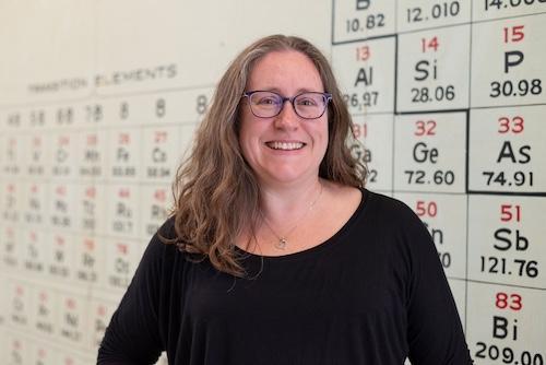 Woman in a black shirt wearing glasses and smiling at the camera as she stands in front of a whiteboard with the periodic table