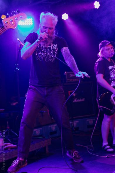 A musician is performing on stage, singing into a microphone. The scene is illuminated with purple and blue lights, creating a vibrant atmosphere. Another band member can be seen in the background, playing a guitar. Amplifiers are visible behind them.