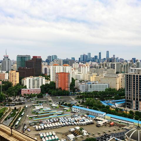 Aerial shot, a parking lot for trucks and a bus terminal in the foreground, residential highrises in the center, and the CBD in the background under a cloudy sky.