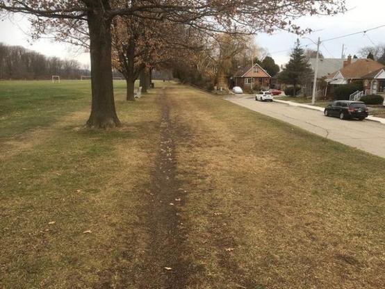 A dirt path lined with trees leads towards a residential area. On the right, there are houses and parked cars, while a soccer field is visible in the background. 