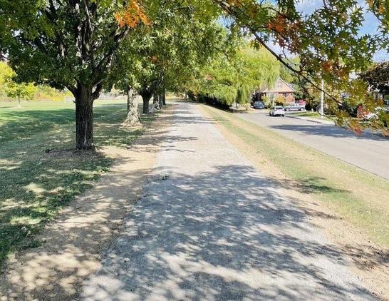 A gravel pathway lined with trees, where some leaves show signs of fall colors. On the right, a street with parked cars, and a grassy area is visible on the left.