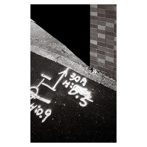 A sharply angled corner creates a striking visual divide between textured tile, dark shadow, and sunlit asphalt. Painted construction markings—arrows, numbers, and symbols—are scrawled across the pavement in white spray, looking both urgent and cryptic.