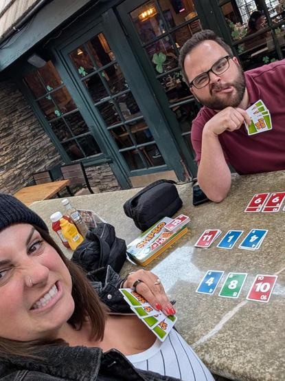 AJMaxwell making a silly face in the background resting his head on his hand that is holding Skip-Bo cards. AJMaxwell's wife is in foreground also making a silly face. Skip-Bo cards, partially finished beer glasses, and empty shot glasses sit between them.