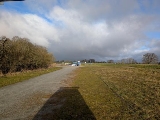 Wir stehen neben einem geschotterten Weg und ich knipse hinten aus dem Camper heraus. Am Ende des Weges ist ein Seitenarm der Elbe, der im Sommer als Sportboothafen fungiert. Auf dem Wasser ist ein schwimmendes Café, das aber zurzeit noch geschlossen ist. Rechts von uns ist der Deich. Der Himmel ist heiter bis wolkig.