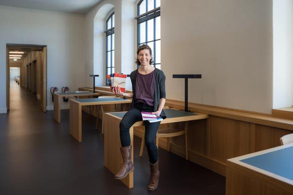 Auf dem Bild sieht man einige Arbeitsplätze in der Universitätsbibliothek Chemnitz. An einem Tisch lehnt Friederike Kimme, eine Mitarbeitende im Universitätsverlag Chemnitz. Sie hält einige Printexemplare von Publikationen in den Händen, die im Universitätsverlag erschienen sind. 