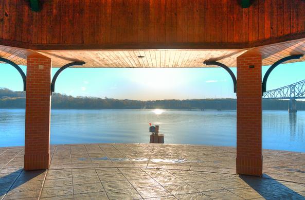 A riverside gazebo frames the view of a tranquil Mississippi River under a clear sky, with the sun rising near the horizon.