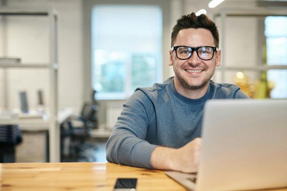 A smiling man wearing glasses is using a laptop, showcasing a positive and engaged expression