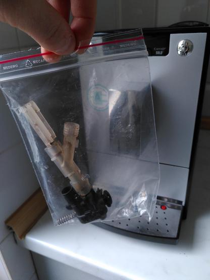 Kaffeemaschine auf einer Arbeitsplatte der Küche. Davor eine Tüte gehalten mit Plastikteilen und Feder eines Drainageventils.
